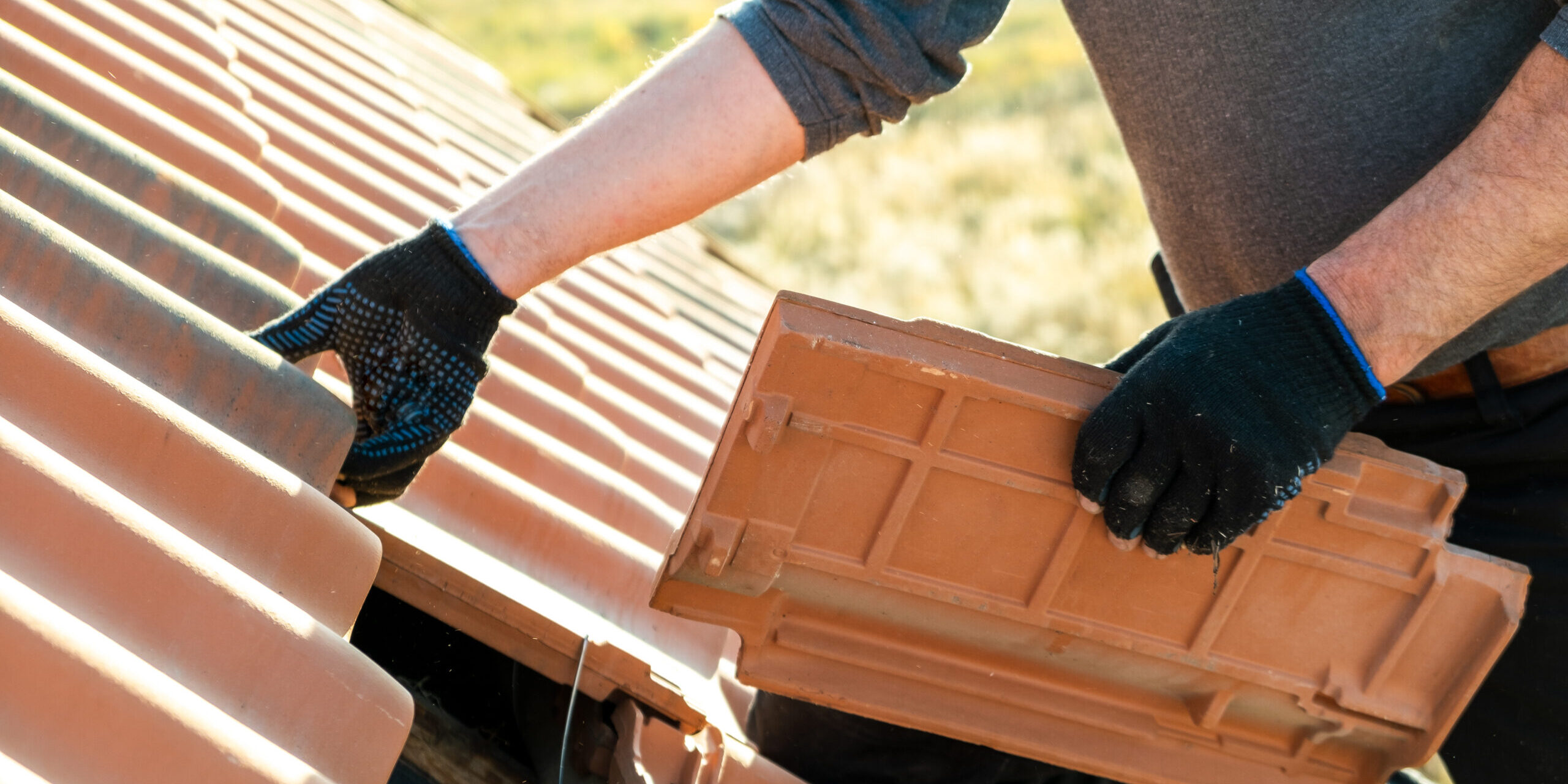 Closeup of worker hands installing yellow ceramic roofing tiles mounted on wooden boards covering residential building roof under construction.